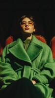 Stylish woman in vibrant green coat and sunglasses seated in an elegant theater with deep shadows photo