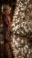Child in cozy pajamas gazes at Christmas tree adorned with twinkling lights during holiday season photo
