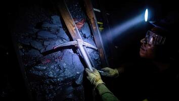 Miner uses pickaxe to extract coal in a dark underground tunnel while illuminated by a headlamp photo