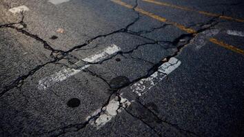 Cracked asphalt road showing signs of wear and age with visible yellow markings and debris in an urban area during daylight hours photo