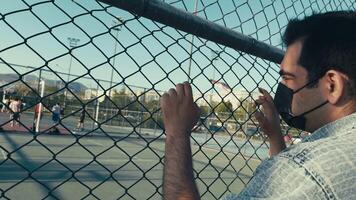 Young Unhealthy Sick Man with Protection Mask Watching Basketball Players in Field Behind the Metallic Fence video