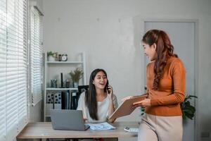Two women working together in a modern office, discussing documents and using a laptop. photo