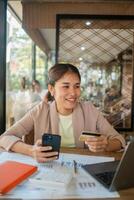 Smiling woman using smartphone and credit card for online shopping in a cafe, with documents and laptop on the table. photo