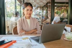 A young woman in a cafe working on a laptop, surrounded by documents and a calculator, showcasing a modern work environment. photo