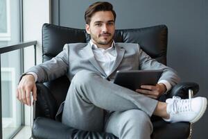 Young, successful entrepreneur in a stylish grey suit and white sneakers, relaxing confidently in a black leather executive chair while holding a tablet, with a large window view in a modern office photo