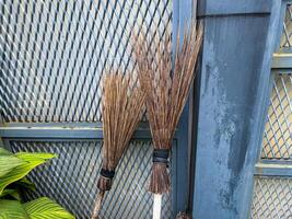 Two brooms are leaning and resting upright in front of a sturdy metal fence, creating an interesting scene in the outdoor area nearby photo