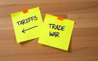 Tariffs reminder two yellow sticky note with the word TARIFFS and TRADE WAR handwritten in black on a wooden surface, Import tariffs increase concept photo