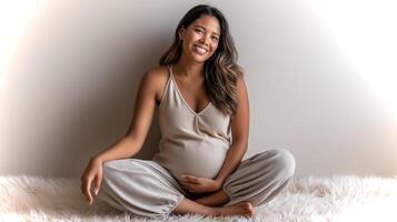 A latina woman with a glowing smile sits comfortably on a soft rug, showcasing her pregnancy while wearing a beige tank top and loose flowy pants. photo