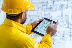 A man in a hard hat is using a tablet to look at a blueprint photo