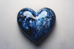 A glossy blue heart resting on a light gray surface, reflecting soft light in an artistic display of color and shape photo