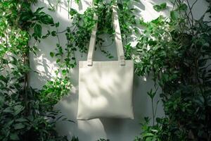 A minimal canvas tote bag hangs among lush green plants in soft natural light, showcasing a blank space for customization photo