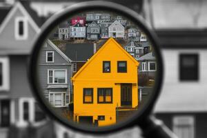 A magnifying glass highlights a striking yellow house set against a backdrop of grey homes in a picturesque neighborhood photo