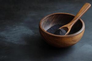 Activated charcoal powder displayed in a wooden bowl with a spoon on a textured black background, emphasizing its natural appeal photo