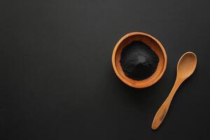 Activated charcoal powder in a wooden bowl with a spoon on a black background for natural health and beauty applications photo
