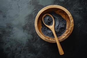 Activated charcoal powder in a wooden bowl with a spoon set against a sleek black background for natural health and beauty remedies photo