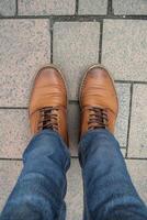 A casual male outfit featuring denim jeans and brown leather shoes while standing on a textured pavement during the day photo