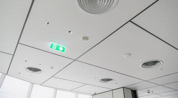 Clean modern office ceiling featuring air vents, recessed lighting, and grid pattern with illuminated emergency exit sign, creating bright and organized atmosphere photo