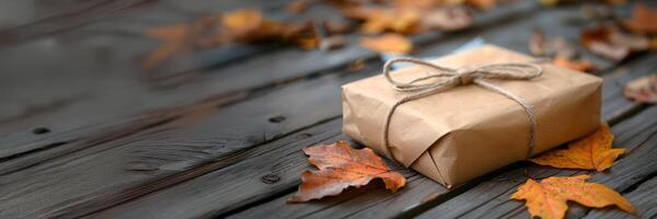 Banner with a gift wrapped in brown kraft paper and tied with a bow placed on a wooden surface surrounded by colorful autumn leaves. Autumn background with copy space for text, advertising and message photo