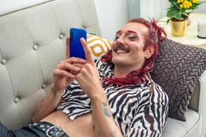 Non-binary person with pink hair using smartphone and smiling on sofa photo