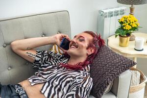 Non-binary person with pink hair talking on phone and smiling, lying on sofa at home photo