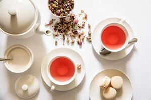 dos tazas lleno con Rosa infundido té sentar junto a delicado macarons y un cuenco de seco flor pétalos en un limpiar blanco mesa. un tetera y cuchara completar el atractivo configuración. foto