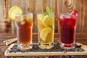 Three tall glasses filled with iced tea, lemonade with lemon slices, and a berry drink with cherries are presented on rustic wooden table. Fresh mint garnish adds vibrant touch. photo