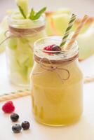 Jar with melon smoothie near berries, straws diagonally behind a jar with melon pieces, melon cut slices on a white background. Melon smoothie and salad in 2 jars. Vertical. Close-up. photo