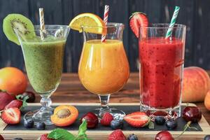 Three tall glasses filled with green, orange, and red smoothies are arranged on a wooden table, surrounded by fresh fruits like strawberries, kiwis, and cherries, perfect for hot days. photo
