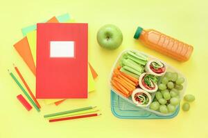 Lunchbox showcases vibrant assortment of fruits and vegetables, including cucumbers, carrots, grapes, and apple alongside wraps. Colorful stationery adds to cheerful presentation. photo