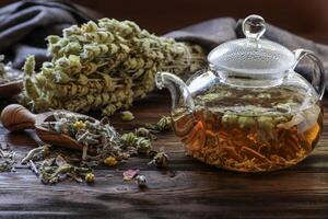 Tea is steeping in glass teapot, surrounded by fresh herbs and dried flowers on weathered wooden table, creating warm and inviting atmosphere for relaxation and enjoyment. photo