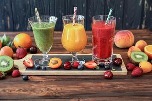Three tall glasses of smoothies in green, yellow, and red hues sit on wooden serving board. Surrounding fruits include strawberries, kiwi, peaches, and oranges, creating a refreshing atmosphere. photo