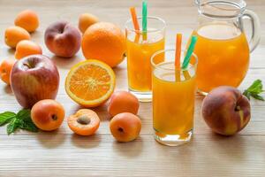 Fruit juice in glasses with ice near the jar of juice and scattered apricots, peaches, apple, orange on a light wooden background. Apricot peach apple orange juice with ice. Horizontal. photo