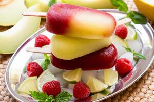 Vibrant melon raspberry ice cream sits atop a mound of ice cubes, surrounded by fresh raspberries, mint leaves, and sliced lime. photo