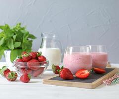 Two glasses of strawberry smoothie placed on serving board next to bowl of fresh strawberries and pitcher of milk, surrounded by mint leaves, creating vibrant atmosphere. photo