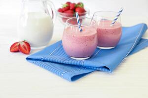 Two colorful strawberry smoothies in clear glasses are placed next to a pitcher of milk and a bowl of fresh strawberries on a blue napkin. photo