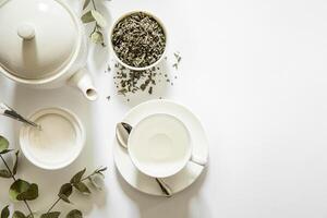 Teapot, cup, and plate arranged neatly with loose herbal tea, sugar bowl, and eucalyptus leaves on white surface, creating serene atmosphere for tea enjoyment. photo