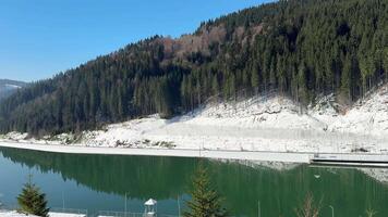 Sunlight illuminates snow-covered shores and evergreen forest surrounding nagold dam reservoir, creating a serene winter scene with reflections on the calm water video
