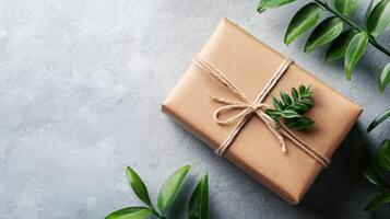 A neatly wrapped gift box with a green leaf on top, placed on a grey stone surface with green leaves in the foreground and a grey background. photo