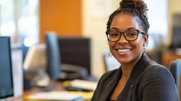 A smiling woman with glasses and a bun, wearing a black blazer, sitting in an office with a computer in the background. photo