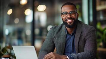 A smiling man with a beard and glasses, wearing a gray sweater, sitting at a desk with a laptop in a modern office setting. photo