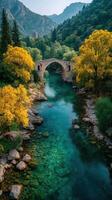 Beautiful autumn landscape featuring a stone bridge over a crystal-clear river surrounded by colorful trees in the mountains photo