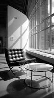 A black and white photo of a chair and table in front of a large window