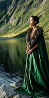 Viking woman in green cloak stands by serene lake surrounded by mountains photo