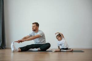 Teaching how to stretch the legs. Father with little daughter are doing yoga at home photo