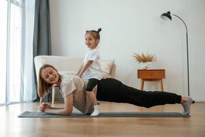 Kid is sitting on a back of woman... Young mother with little girl are doing yoga at home photo