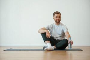 Taking a ,break,, with bottle of water. Yoga man is sitting on a mat at home photo