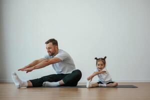 Against ,white wall, ,,stretching. Father with little daughter are doing yoga at home photo