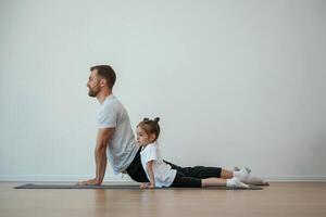 ,Against white wall, stretching. Father with little daughter are doing yoga at home photo