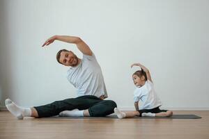 Against white ,wall, stretching. Father with little daughter are doing yoga at home photo