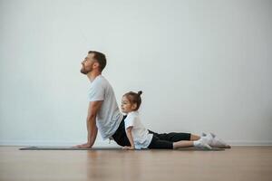 Against, white wall, stretching. Father with little daughter are doing yoga at home photo